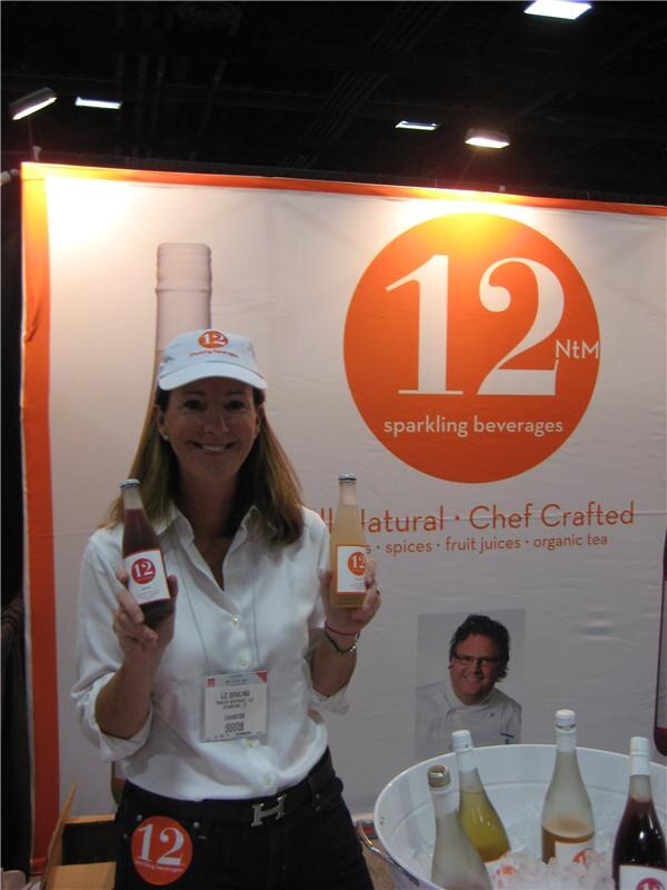 A smiling woman in a white cap and white shirt holds up two bottles in front of a 12 NtM sparkling beverages backdrop. A tub of ice with more bottles is in front of her.