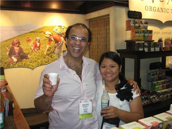 Ahmed Rahim, at left, smiles and holds a small paper cup, standing with a woman holding a bottle at the Numi booth, with tea boxes on shelves behind them.