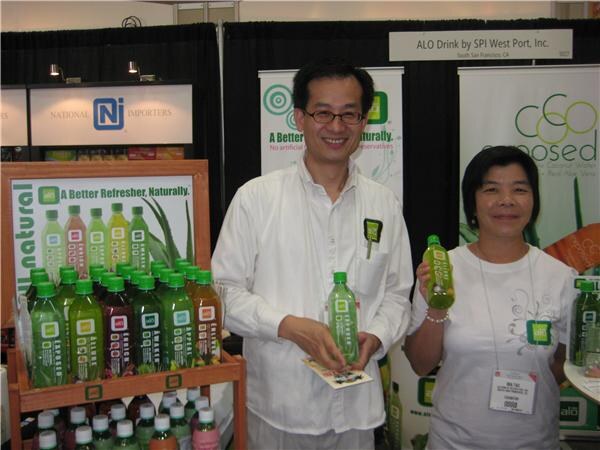 A smiling man in glasses and a white shirt and a smiling woman hold green ALO Drink bottles in front of a shelf of bottles in the ALO Drink by SPI West Port booth, next to National Importers signage.
