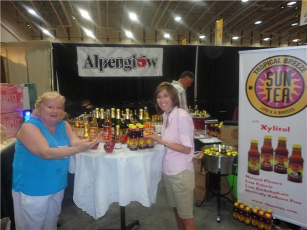 Two women stand at a white-draped table of bottled drinks and red apples beneath an Alpenglow banner, with a Tropical Breeze Sun Tea banner on the right and a man behind them.