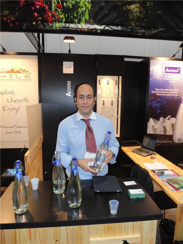 A man in a light blue shirt and red tie holds a glass bottle of Aonni water behind a black counter with more bottles, in a booth marked Aonni with a Chile banner beside it.