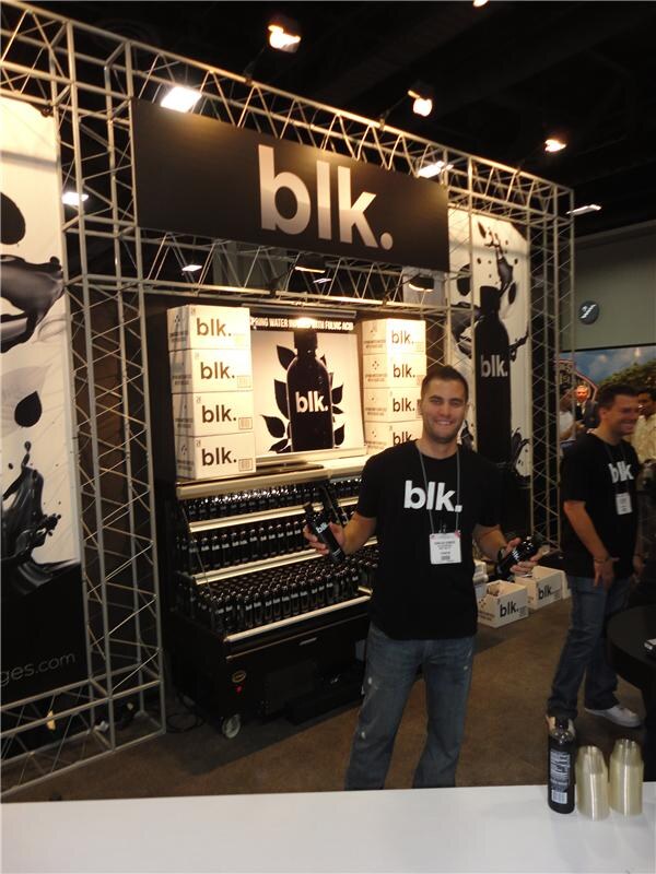 A smiling man in a black blk. T-shirt holds two dark bottles in front of a truss booth with a blk. sign and stacked blk. cartons; another man in a black shirt stands at right.