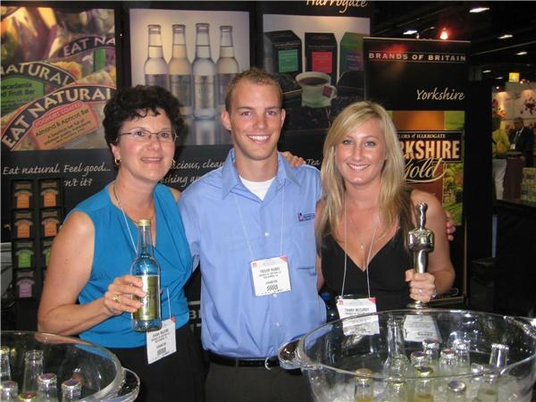 Two women and a man stand together at the Brands of Britain booth with Eat Natural and Yorkshire Gold signage behind them; one woman holds a bottle and the other holds a small trophy.
