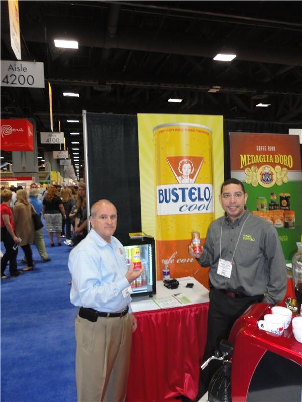 Two men stand at a booth with a tall yellow Bustelo Cool can banner and a Medaglia D'Oro banner; each holds a can, and a red espresso machine is at the right.