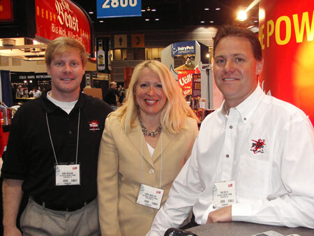 Two men and a blonde woman wearing name badges pose close together in front of a red booth. A DairyPak sign and other trade show signage appear in the background.