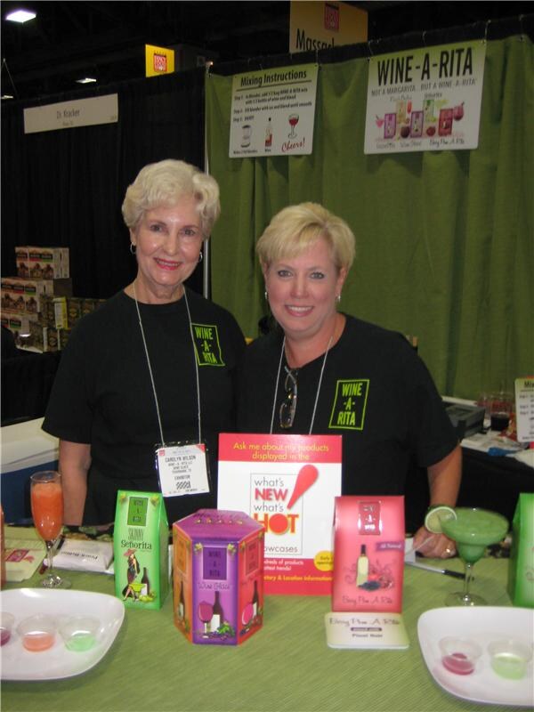 Carolyn Wilson, left, and a woman beside her wear black Wine-A-Rita shirts behind a table of Wine-A-Rita boxes, cocktails in glasses and sample cups, with a green Wine-A-Rita banner behind.