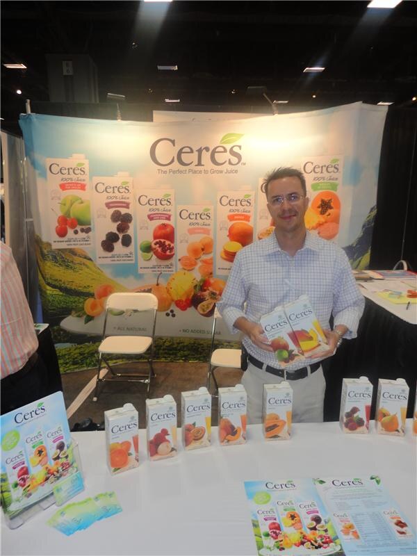 A man in a checked shirt and glasses holds two Ceres juice cartons in front of a Ceres booth backdrop, with more cartons and flyers on the table in front of him.