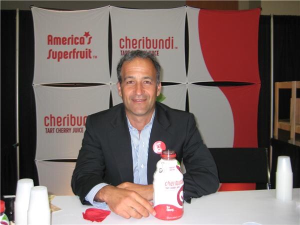 A smiling man in a dark blazer sits at a table with a bottle of Cheribundi tart cherry juice, in front of a banner reading America's Superfruit and Cheribundi.