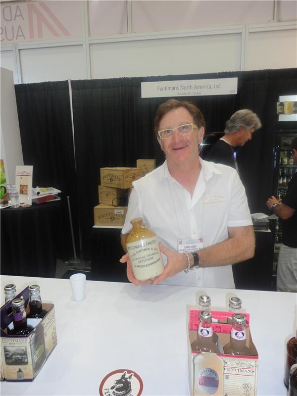 Craig James, in glasses and a white shirt, holds a stoneware Fentimans bottle at the Fentimans North America, Inc. booth, with boxed bottles of Fentimans drinks on the counter.