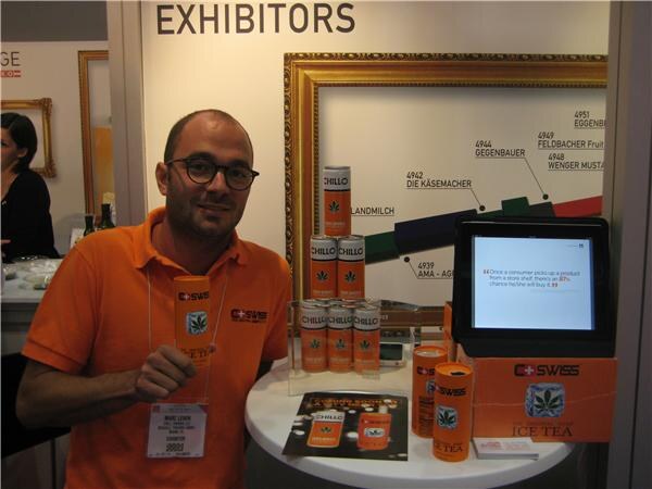 A smiling man in glasses and an orange C-Swiss shirt holds a can of Chillo ice tea beside a table of Chillo cans and a tablet screen.