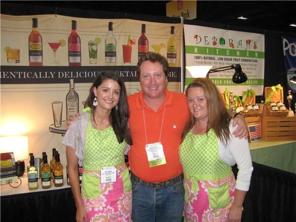 A man in an orange polo stands between two smiling women in green and pink aprons at the Deborah's Kitchen booth, with cocktail mixer bottles and banners behind them.