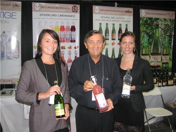 A man stands between two women, each holding a bottle, in front of Epicurean Beverages banners for sparkling limonades and sparkling juices.