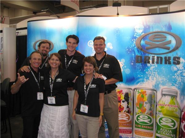Five people in black Ex Drinks shirts and badge lanyards pose together in front of an Ex Drinks backdrop showing cans and a bottle of Aqua Vitamins.
