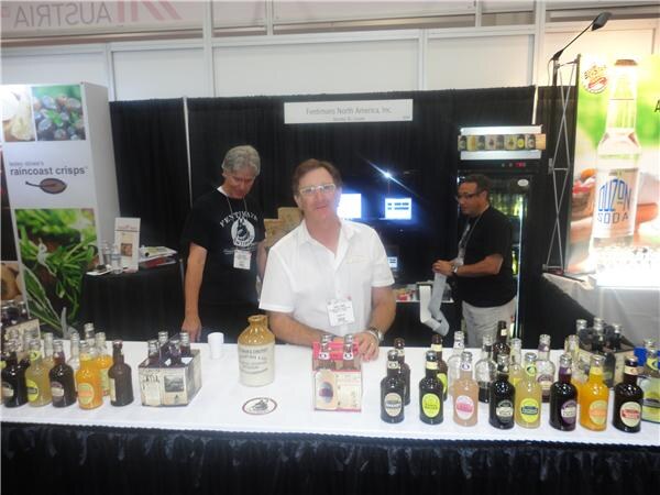 A man in a white shirt stands behind a counter of Fentimans bottles at the Fentimans North America, Inc. booth, with two other people behind him and a Raincoast Crisps banner at left.