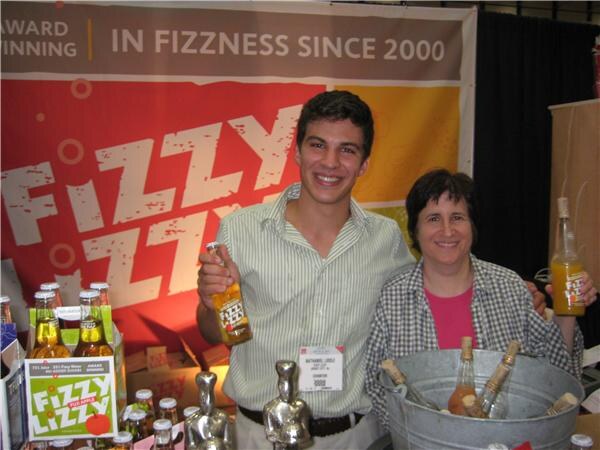 A young man holding a Fizzy Lizzy bottle stands next to a smiling woman with a bucket of bottles, in front of a Fizzy Lizzy banner reading In Fizzness Since 2000.