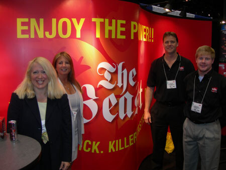 Two women and two men pose in front of a red The Beast backdrop reading 'Enjoy the power!!'. Two cans sit on a small table at left, and the men wear black shirts and name badges.