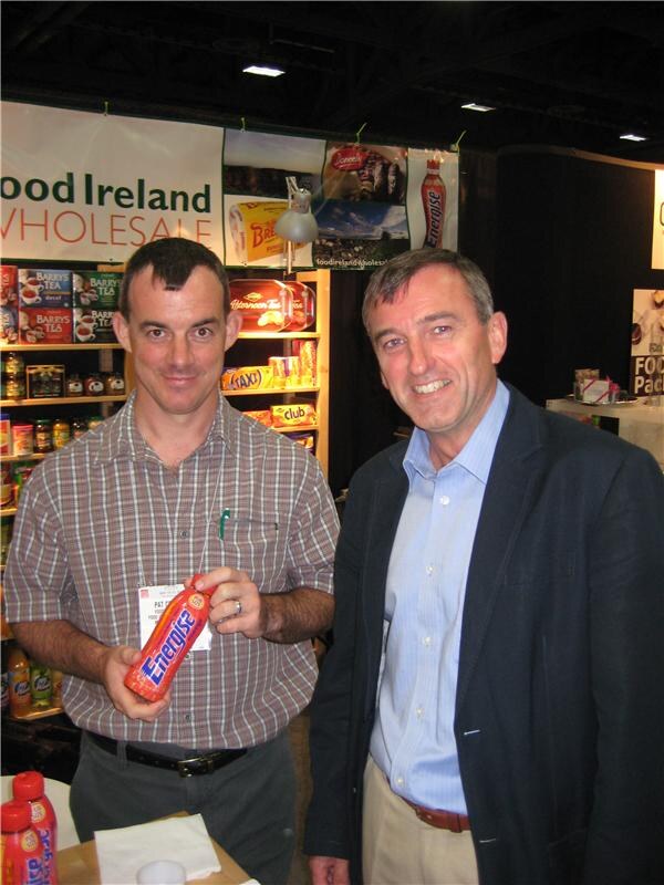 Two smiling men stand at the Food Ireland Wholesale booth; one holds a bottle of Energise, with Barry's Tea boxes on shelves behind them.