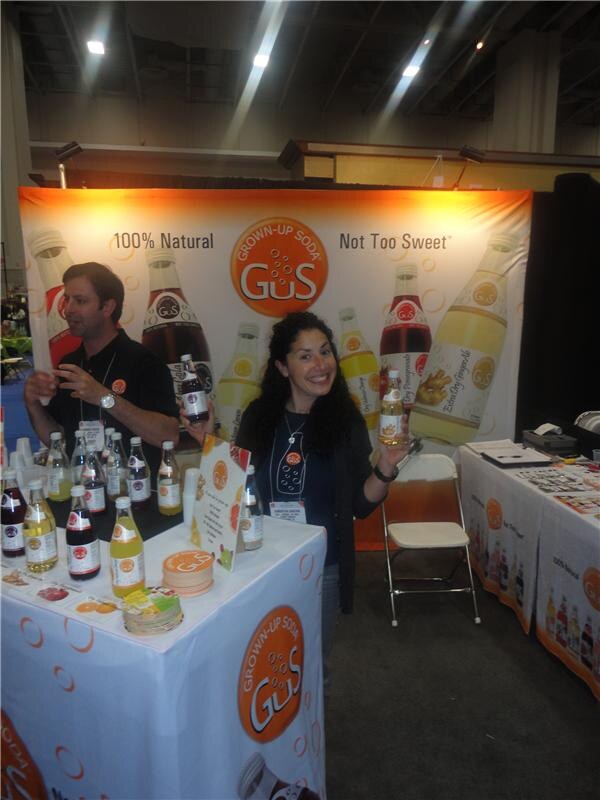 A smiling woman holding a bottle and a man in a black shirt stand at a table of GuS Grown-Up Soda bottles, in front of a GuS banner reading 100% Natural, Not Too Sweet.