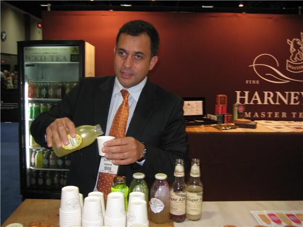 A man in a dark suit and orange tie pours tea from a bottle into a paper cup at the Harney & Sons booth. Iced tea bottles are in a cooler behind him and ginger ale bottles are on the counter.