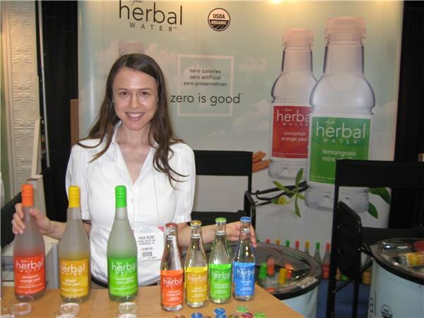 A smiling woman with long brown hair stands behind a table of Herbal Water bottles in several colors, in front of a banner reading zero is good.