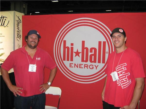 Two men in red hi-ball shirts and caps stand with hands on hips in front of a large red hi-ball Energy sign, both wearing badges on lanyards.