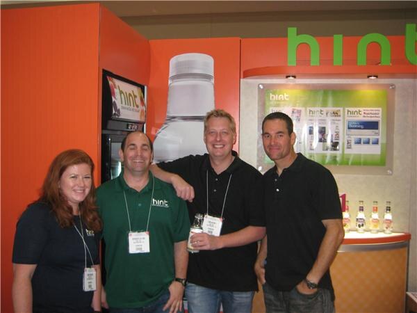 A woman and three men pose together at the orange Hint booth. One man holds a small bottle, and a large bottle graphic and a shelf of bottles are visible behind them.