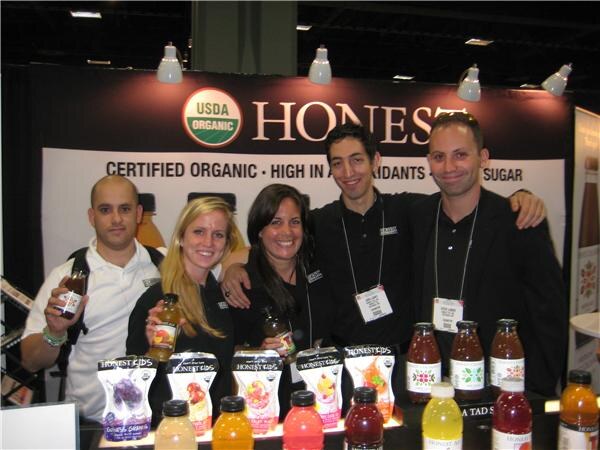 Five people smile and pose behind a counter of Honest Kids pouches and bottled drinks, some holding bottles, in front of a Honest USDA Organic sign.