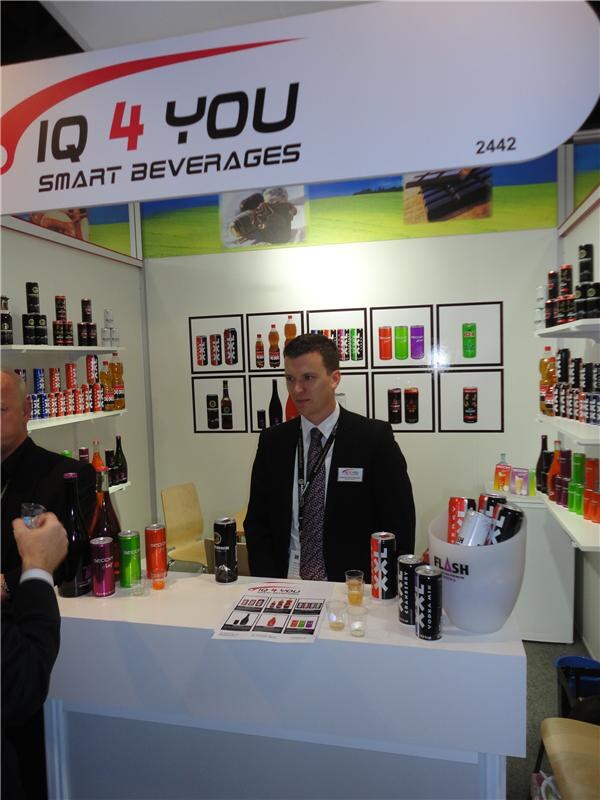 A man in a dark suit stands behind a white counter at the IQ 4 You Smart Beverages booth, with energy drink cans, a Flash ice bucket, small cups and shelves of bottles around him.
