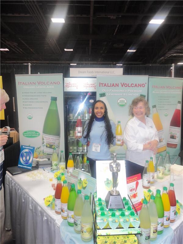 Two women stand behind a table of Italian Volcano lemon juice, lemonade and limeade bottles, with Italian Volcano banners and a sign reading Dream Foods International LLC. behind them.