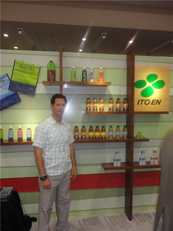 A man in a checked short-sleeved shirt stands before wooden shelves of bottled Ito En teas beside a lit Ito En sign. A Teas' Tea bag hangs on the left.