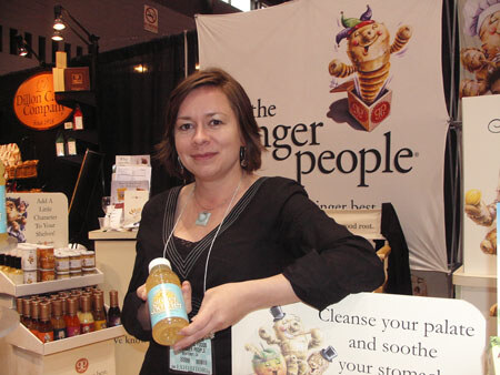 A smiling woman with dark hair holds a bottle of yellow drink at The Ginger People booth, with The Dijon Cart Company sign and shelves of bottles behind her.