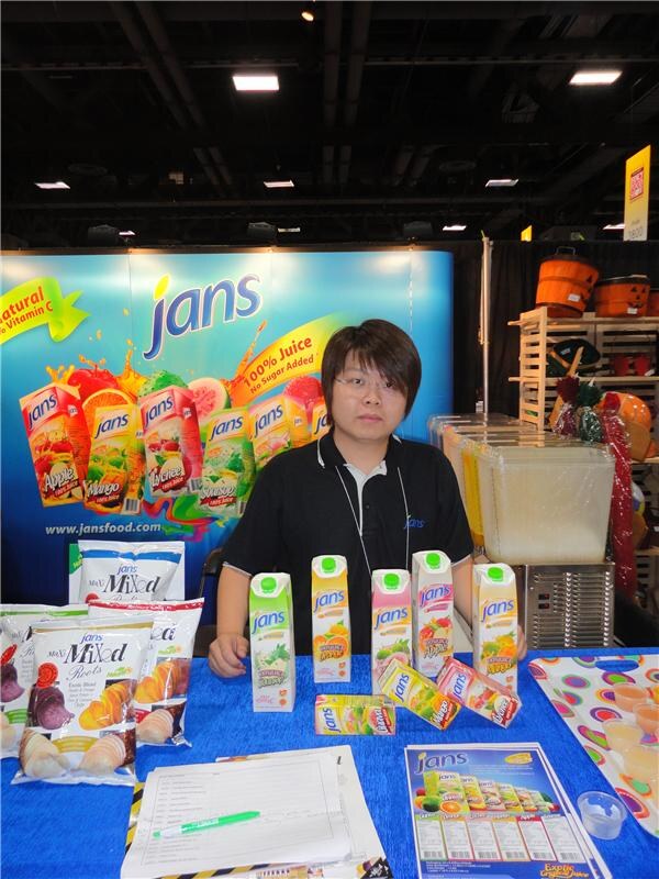 A person in a black shirt sits behind a blue-covered table at the Jans booth, with juice cartons, bags of chips and printed sheets in front of a Jans juice backdrop.