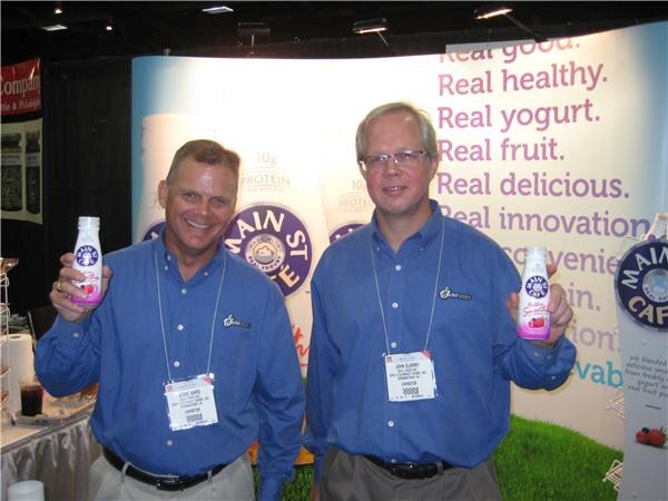 Two men in blue shirts and exhibitor badges each hold a bottle of Main St Café drink in front of a Main St Café banner reading Real good, Real healthy, Real yogurt.