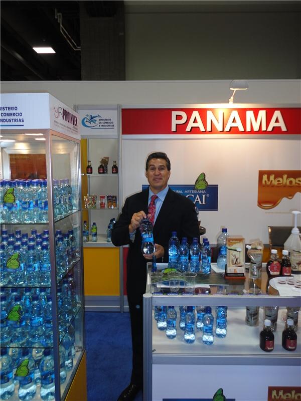 A smiling man in a dark suit holds a bottle of water at a booth under a red Panama sign, with bottled water in display cases and Melos signage visible.