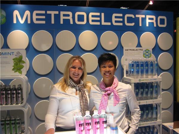 Two smiling women in white shirts and scarves stand behind bottles of pink-labeled water at the Metroelectro booth, with shelves of bottled water behind them.