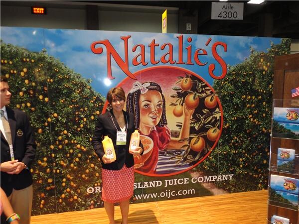 A woman in a dark jacket and red patterned skirt holds two juice jugs in front of a large Natalie's Orchid Island Juice Company backdrop of orange groves. A man stands at left.