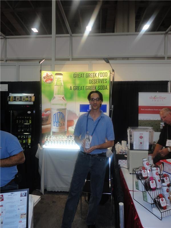 A man in a blue polo and glasses holds a bottle of Ouzon Soda in front of a banner reading Great Greek food deserves a great Greek soda. A Shoreline Fruit sign is at right.