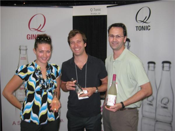 A woman and two men stand in front of Q Tonic banners. The woman and the man on the right each hold a bottle, and the man in the middle holds a small boxed item.