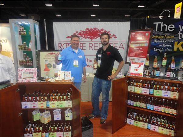 Two men stand in the Rooibee Red Tea booth between open wooden cabinets filled with bottled tea. A banner behind them has the Rooibee Red Tea name and an Orthodox Union sign.