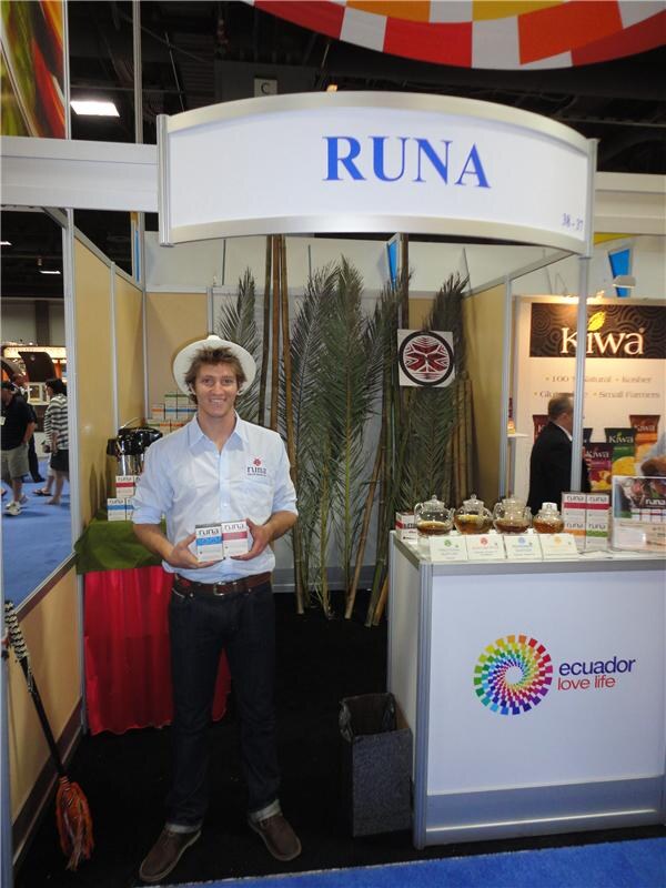 A smiling man in a white hat and shirt holds two boxes of tea at the RUNA booth. Teapots sit on the counter, and Kiwa and Ecuador Love Life signs appear nearby.