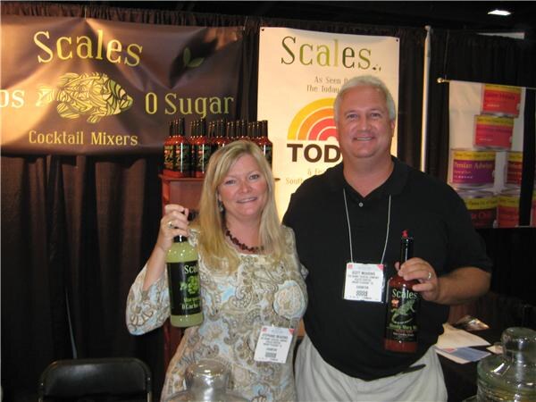 A blonde woman and a man in a black shirt each hold a bottle of Scales cocktail mixer. Scales banners, one with a Today show logo, hang behind them.