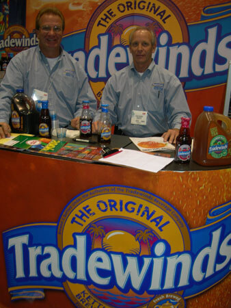 Two men in gray button-up shirts sit behind a Tradewinds counter with bottles of iced tea, brochures and papers on top. A large jug of Tradewinds tea stands at right.