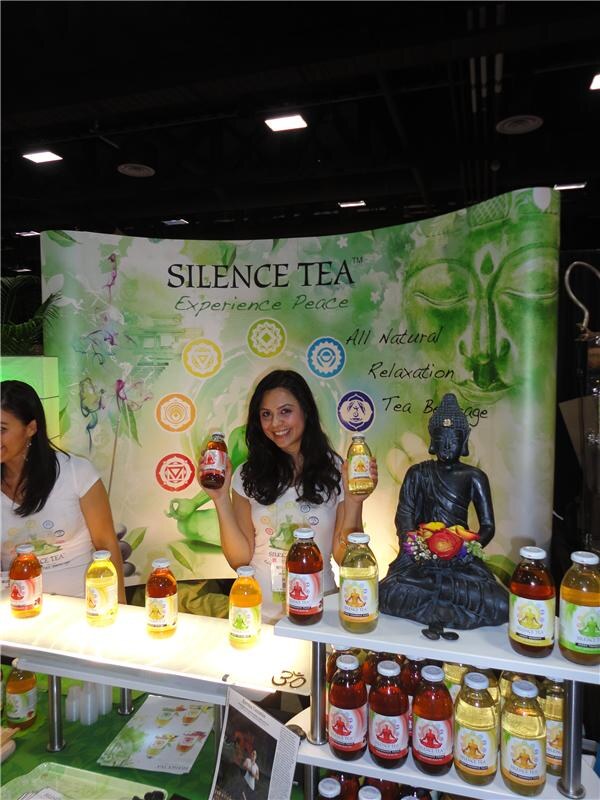 A smiling woman holds up two bottles of Silence Tea in front of a green banner with a Buddha face. A Buddha statue and rows of bottled tea sit on shelves beside her.