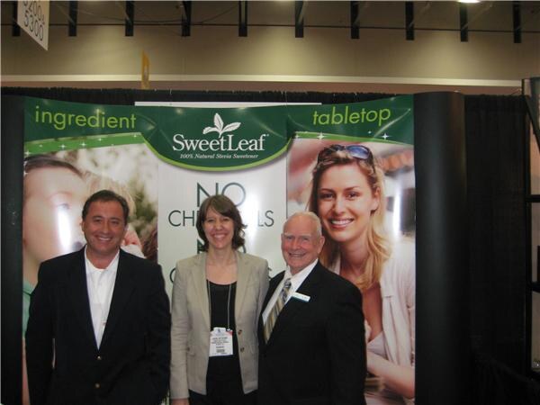 A man in a dark jacket, a woman in a beige blazer and an older man in a suit stand in front of a green SweetLeaf banner showing the words ingredient and tabletop.