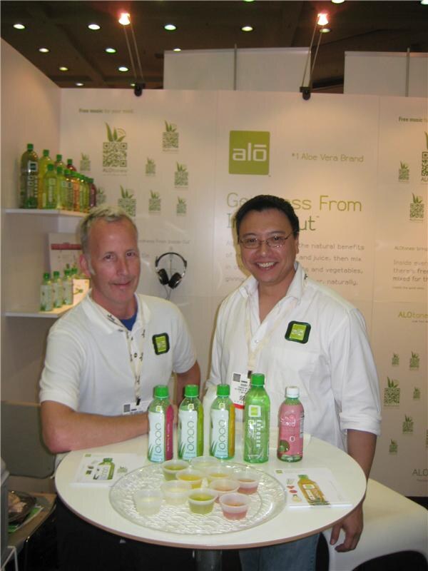 Two men in white shirts stand behind a round table holding bottles of Alo aloe vera drink and small sample cups, in front of Alo branded wall graphics and a shelf of bottles.