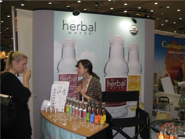 A woman in a leopard-print top stands at an Ayala's Herbal Water booth with bottles and sample cups on the counter while a woman in black sips a sample. A Carrington sign is behind them.
