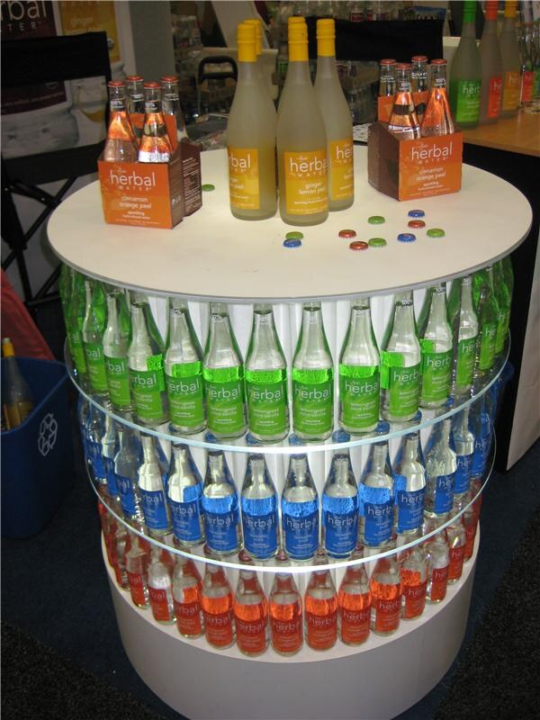 Round white tiered display of Ayala's Herbal Water glass bottles with green, blue and orange labels, topped with ginger lemon bottles, cinnamon orange peel four-packs and scattered bottle caps.