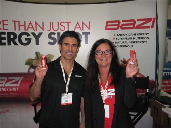 A smiling man and a woman with glasses, both with lanyards, each hold up a small red Bazi energy shot bottle in front of a Bazi banner.