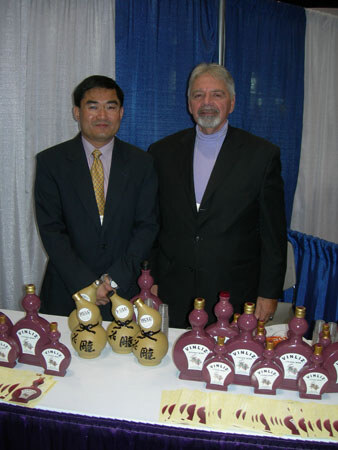 Two men in dark jackets stand behind a table displaying purple Vinliz bottles and several tan decorated bottles, with blue curtains behind them.