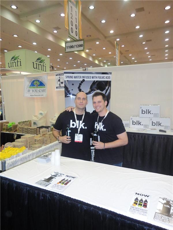 Two men in black blk. T-shirts hold bottles of blk. water behind a white-covered table, with stacked blk. cases, a UNFI sign and an If You Care sign behind them.